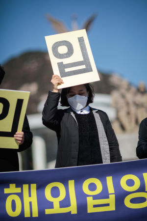 Tillsammans bildar demonstranterna orden: Stoppa internationell adoption omedelbart. En kvinna med munskydd håller upp en skylt med koreanska tecken.