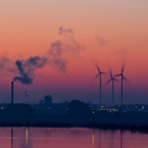 Anläggningar för fossilt bränsle och vindkraftverk vid vatten i solnedgång.