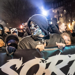 Fyra personer, två av dem maskerade, håller en stor banderoll. Den är svart med vit text. Bakom dem ser man flera människor stå.