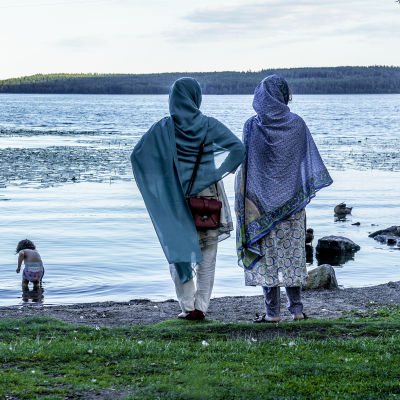 Två kvinnor i slöja står på en sjöstrand och övervakar ett litet barn som plaskar i vattnet.