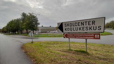 En vit vägskylt med texten Skolcenter Koulukeskus, och en brun skylt med texten Pargas hembygdsmuseum, i en vägkorsning med en skolbyggnad i bakgrunden.