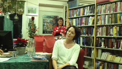 Två kvinnor framför bokhyllor, den ena sitter och den andra står bakom. Till vänster syns ett bord med grön duk, i bakgrunden en julgran.