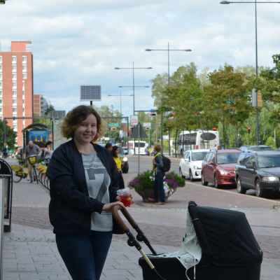 Kvinna står utomhus och framför sig har hon en barnvagn. Hon ler mot kameran och i sin vänstra hand håller hon en plastmugg med sugrör som innehåller en dryck.