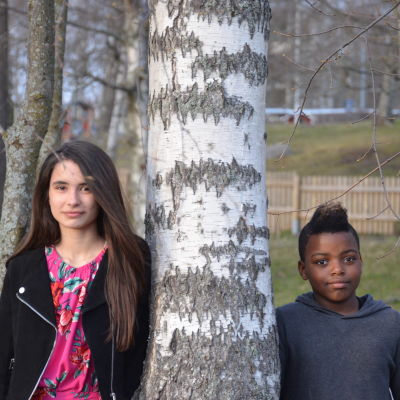 Asta och Valter står på varsin sida av ett träd och ser in i kameran.