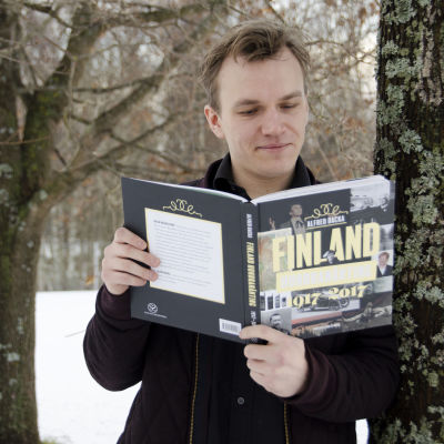 Alfred Backa som står luta mot ett träd och läser sin bok "Finland hundranånting".
