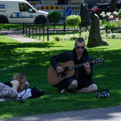 En man samlar in pengar genom att spela gitarr på en gräsmatta på sommaren.