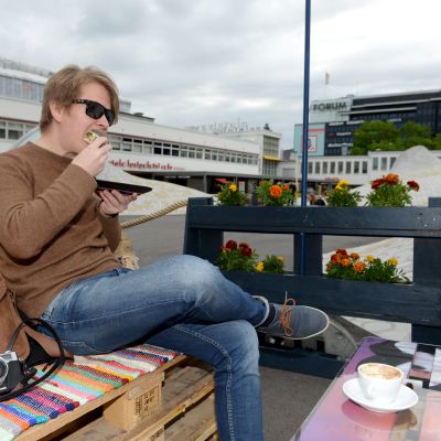 En ung man sitter på ett café i centrala Helsingfors och slukar en smörgås. Framför sig har han en kaffekopp och på bänken ligger han sin läderväska och en kamera,