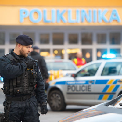 En polis i svarta kläder tittar ner mot sin axel. I bakgrunden finns polisbilar med sirener och ett sjukhus.
