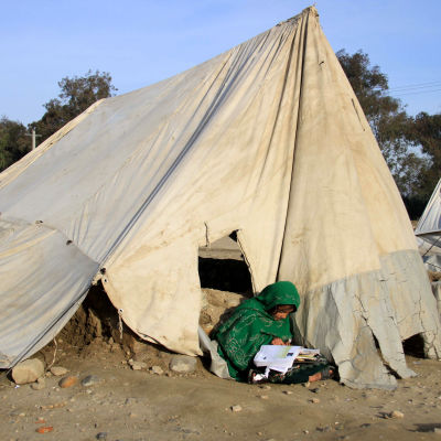 En flicka sitter och läser utanför ett tält. Tältet är gjort av presenning som har spänts mot marken. Det är smutsigt och har hål.