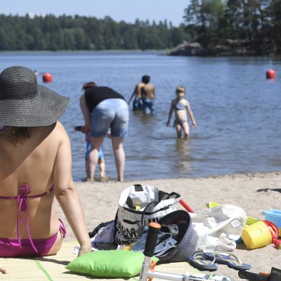 En kvinna sitter på en badstrand i bikini och hatt, i bakgrunden simmar barn och vuxna.