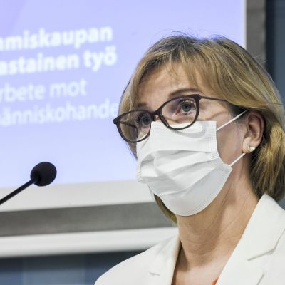 Anna-Maja Henriksson bär munskydd och glasögon. Bakom henne syns en presentation av en handlingsplan.
