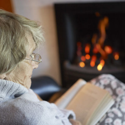 En äldre kvinna sitter och läser en bok framför en öppen spis.