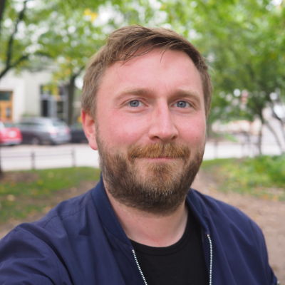 Man i blå skjorta står i en park i Helsingfors och ser mot kameran.