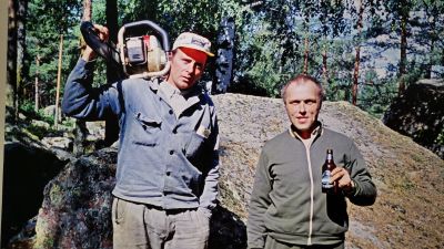 Gammal bild av två män, den ena med en motorsåg på axeln, den andra med en öl i handen. Antagligen på Södra åsen i Lovisa. Aatos Åkerbloms foto.