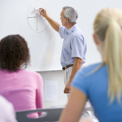 En äldre manlig lärare skriver på en whiteboard i ett klassrum med elever i förgrunden av bilden.