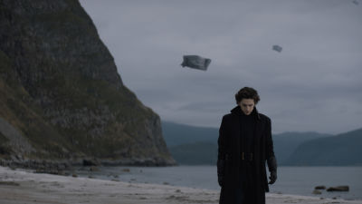 Timothée Chalamet i svart slängkappa på en öde strand.