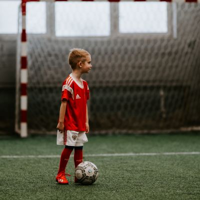 Ett barn står med en fotboll framför sig på en fotbollsplan inomhus.