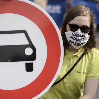 En demonstrant med ansiktsmask håller upp en förbudsskylt mot bilar.