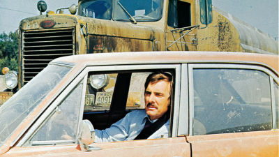 Etualalla mies istuu henkilöauton ratissa. Taustalla näkyy suuri keltainen rekka. Kuva elokuvasta Kauhun kilometrit.