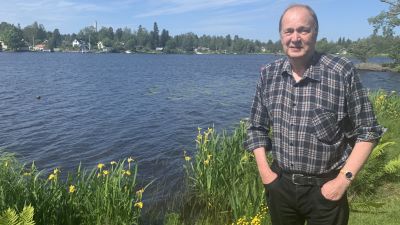 En man i rutig skjorta står med händerna i fickorna vid en sjöstrand.