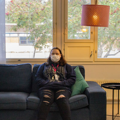 En kvinna med munskydd sitter på en soffa. Hon håller händerna i munktröjans fickor och tittar in i kameran.