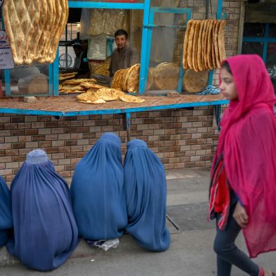 En man säljer bröd genom en liten lucka. Kvinnor klädda i blått sitter på trottoaren framför och en kvinna vandrar förbi.