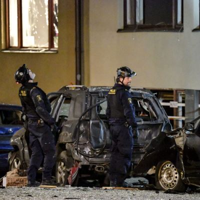 Två män i polisuniformer står på en gata eller en gård framför bilar skadade av en explosion.  Kriminaltekniker arbetar på platsen efter den kraftig explosionen som inträffade i under natten mot måndagen på Östermalm i Stockholm den 13 januari 2020..