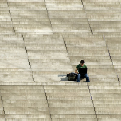 En man som sitter på en stor stentrappa, utomhus i solen,  med en laptop i famnen.