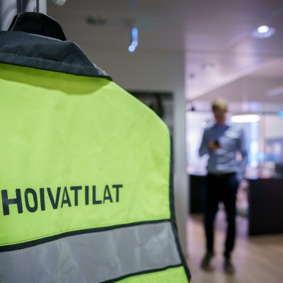 Suomen hoivatilat Oyj:n logo turvaliivin selässä naulakossa. Taustalla epäterävä mieshahmo.