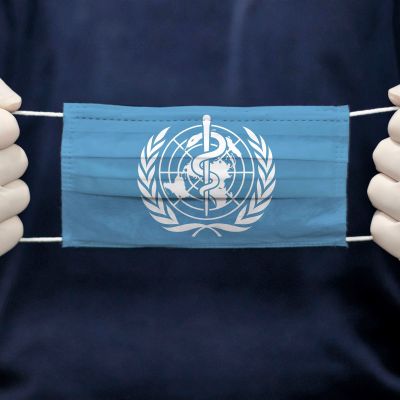 Valkoisiin suojahansikkaisiin puetut kädet pitelevät kasvosuojainta, jossa on sinisellä pohjalla Maailman terveysjärjestön WHO:n valkoinen logo.