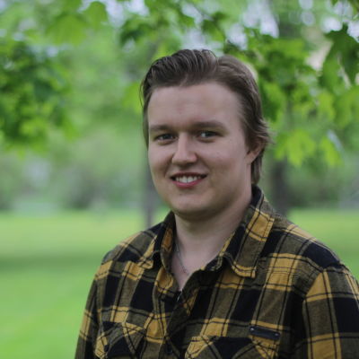 Ung vit man med svart och gulrutig skjorta står framför träd.