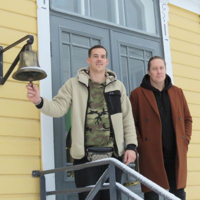 Porträttbild på Nico Rönnberg och Andreas Rönnberg utanför Karis stationshus, ett gult trähus. Nico håller i en stationsklocka som hänger från husväggen. Vinter och snö.