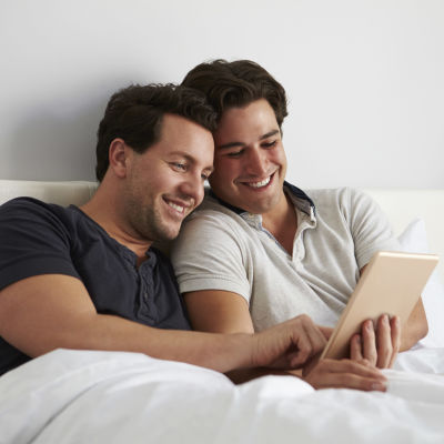 Två leende män ligger i en säng och tittar på en pekskärm