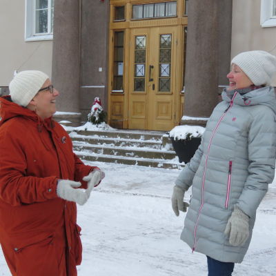 Två kvinnor står utanför Raseborgs stadshus (Ekåsen) vända mot varandra. De skrattar. Vinter och snör. Båda har mössor, vantar och varma vinterrrockar.