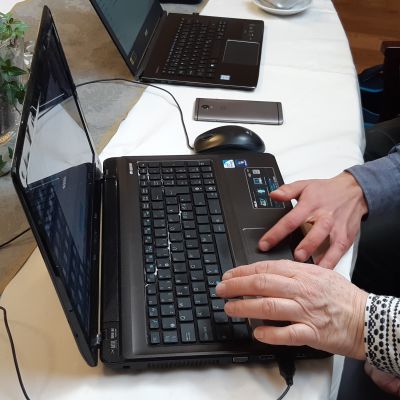 en äldre och en yngre hand ligger på tangenterna till en svart dator. En annan dator syns i bakgrunden och lite av en grönväxt