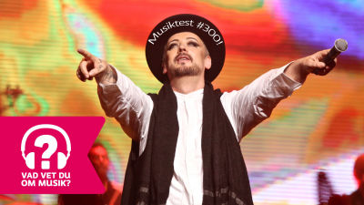 Boy George står med armarna utsträckta framför sig. Han har en mikrofon i vänster hand.