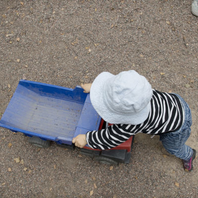 Litet barn i randiga kläder och ljusblå mössa leker med en stor leksaksbil på ett underlag av sand.
