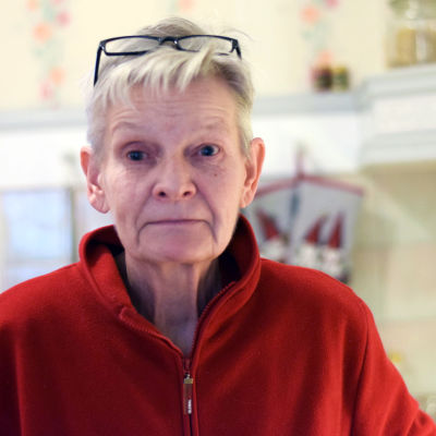 En äldre kvinna med röd fleecetröja och glasögon uppskuffade uppe på huvudet. Hon är i sitt kök.