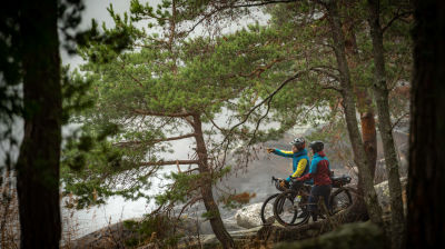 Två cyklister vid en strand med klippor och småväxta tallar. Den ena pekar ut mot havet. Båda bär cykelhjälm.