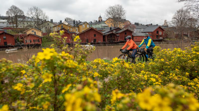 Två cyklister far fram längs en väg invid en å.  Röda hus på andra sidan av ån och gula blommor på samma sida som cyklisterna.