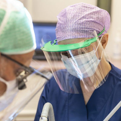 Tandläkare och tandskötare bär munskydd och visir då de jobbar med en patient. 