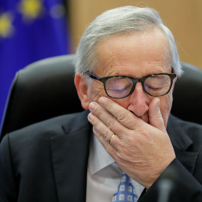 Jean-Claude Juncker silmät kiinni ja käsi suunsa edessä EU-lippu taustallaan.