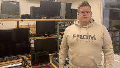 En man med glasögon och munkjacka står intill ett kontrollbord i en tv-studio.