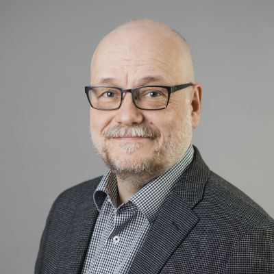 Tunnhårig man med kortklippt, grått skägg, glasögon och mörkgrå, rutig blazer.