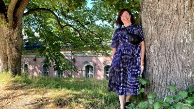 En kvinna med axellångt mörkt hår står lutad mot ett träd. Hon har på sig en blå och vit-prickig klänning, och ser in i kameran och ler.
