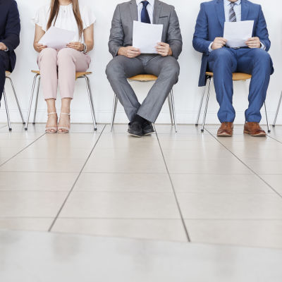 fem personer varav två är kvinnor sitter på stolar och väntar på en arbetsintervju. på bilden figurerar enbart kandidaternas ben.