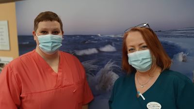 Två hälsovårdare med munskydd framför en tavla med vinterlandskap, Camilla Rautanen i röd sköterskedräkt och Carola Silander-Lehtinen i blå sköterskedräkt.