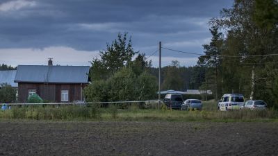 Ett trähus och parkerade bildar syns från en åker. Bland bilarna finns också en polisbil.