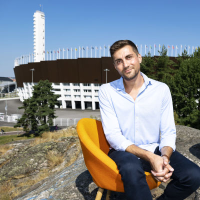 Mies istuu kalliolla oranssilla tuolilla, taustalla näkyy olympiastadion ja toinen oranssi tuoli.
