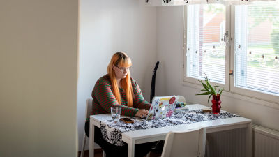 En kvinna med orange hår sitter och jobbar på en dator vid ett vitt köksbord.